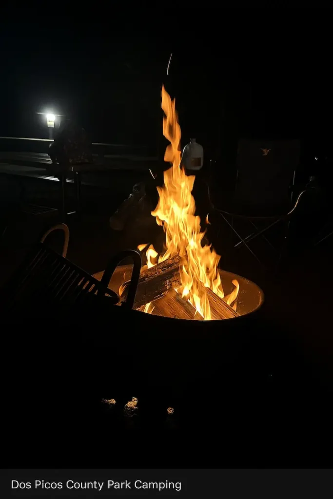 A glowing campfire burns in a metal fire ring at night, surrounded by camping gear including a folding chair, picnic table, and lantern at Dos Picos County Park