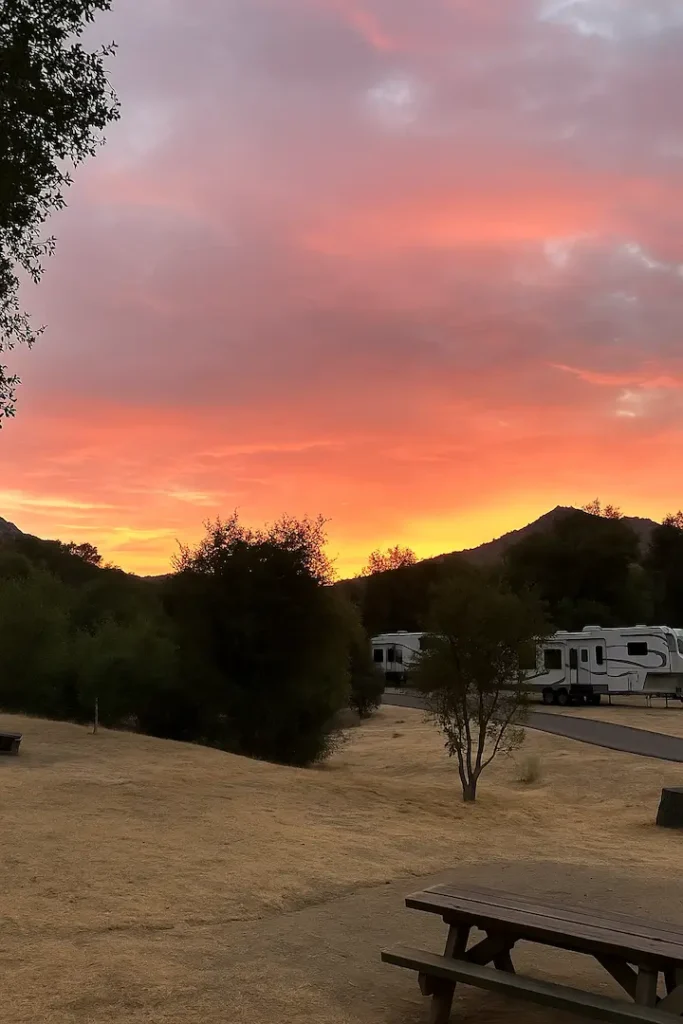 A vibrant sunset lights up the sky over Dos Picos County Park, with RVs parked near a curving road and dry grassy terrain surrounded by trees and hills